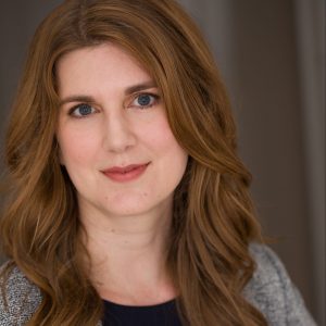 A headshot picture of a white woman with long brown hair wearing a grey sweater over a black shirt.